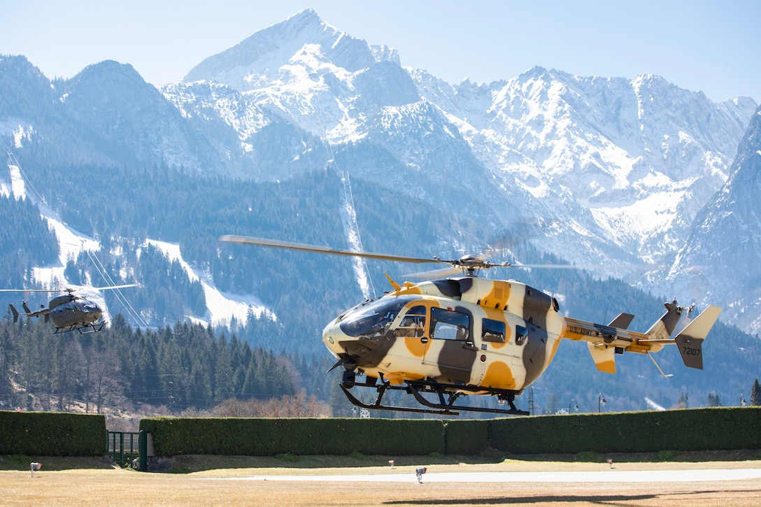 Two helicopters prepare to land with large mountains behind them.