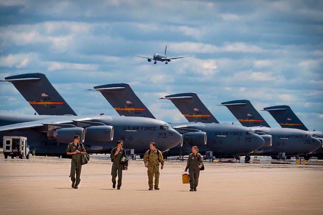 Four airmen walk on a flightline in front or a row of parked aircraft, as a plane flies overhead.