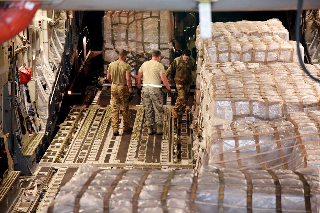 Service members in the cargo hold of an aircraft work with pallets that are wrapped in plastic and strapped down.