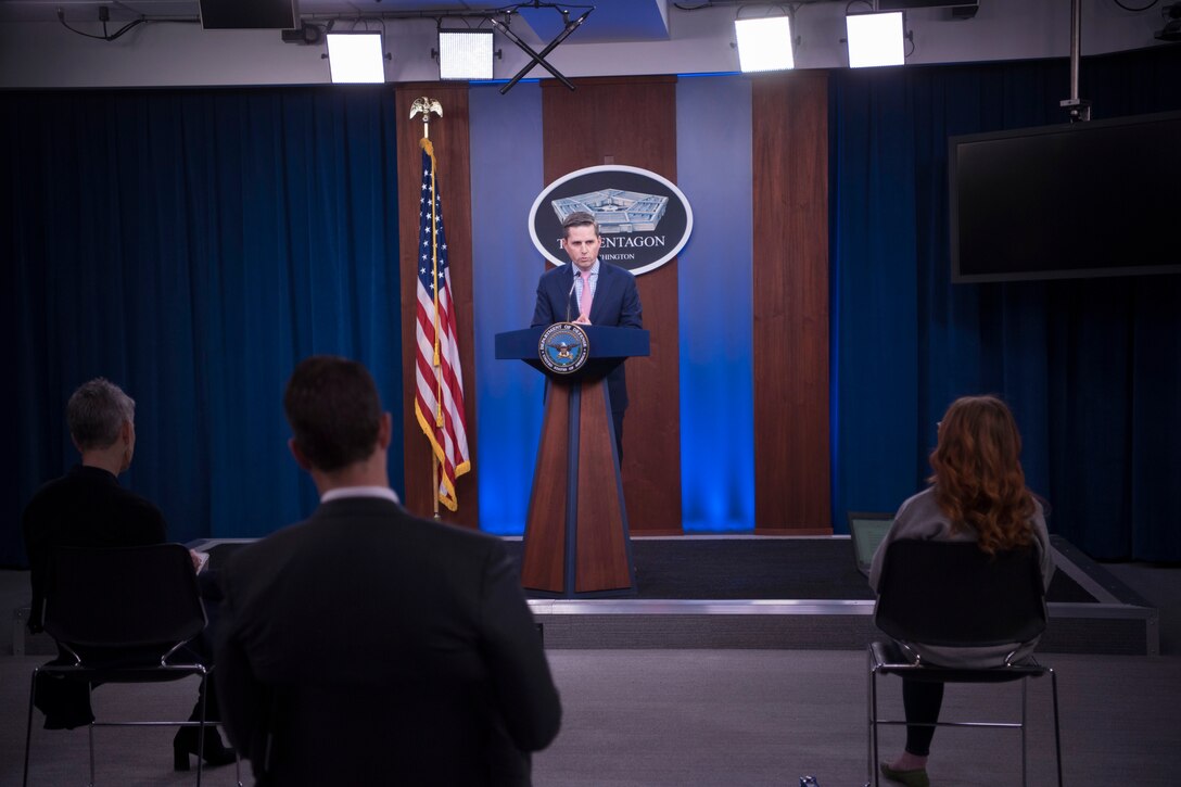 A civilian speaks at a lectern in front of a seated audience.