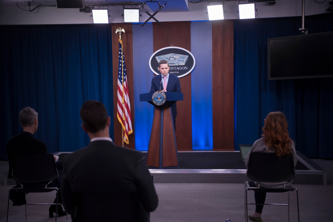 A civilian speaks at a lectern in front of a seated audience.