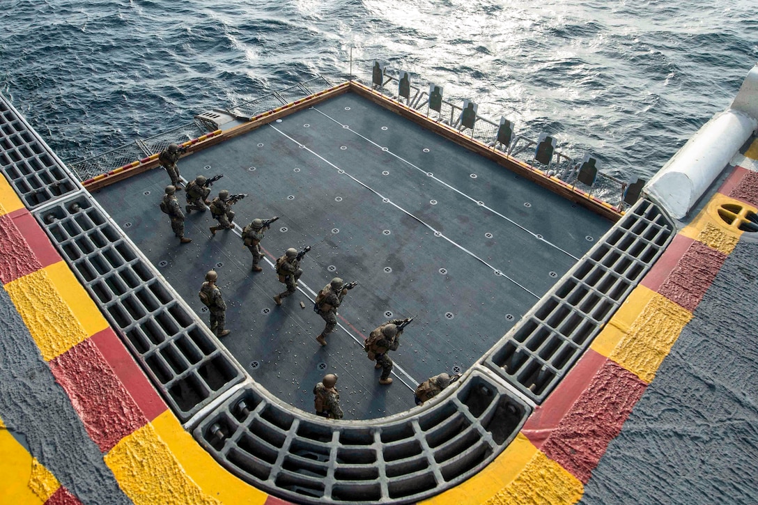 A row of Marines, seen from overhead, fire at targets on a ship deck.