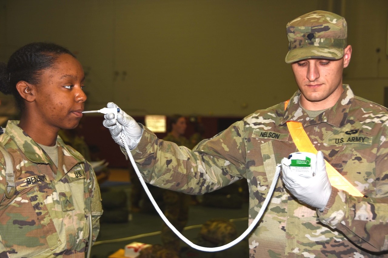 A male soldier in fatigues takes the temperature of a female soldier, who is also dressed in fatigues.