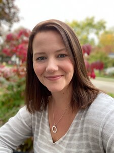 white female with shoulder brown hair in grey and white striped shirt sits outside.