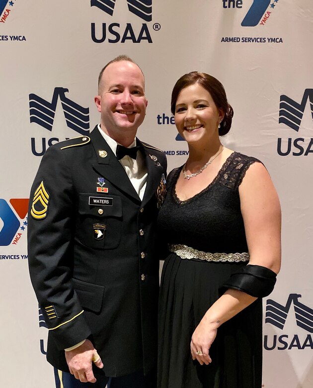 White male in dress uniform stands with white female in black dress with silver belt in front of white background with blue and red and blue eagles that say USAA.