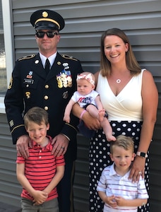 White male in dark dress uniform with hat, white female in white and black dress with polka dots holding a baby with a hairbow and two young boys in striped shirts.