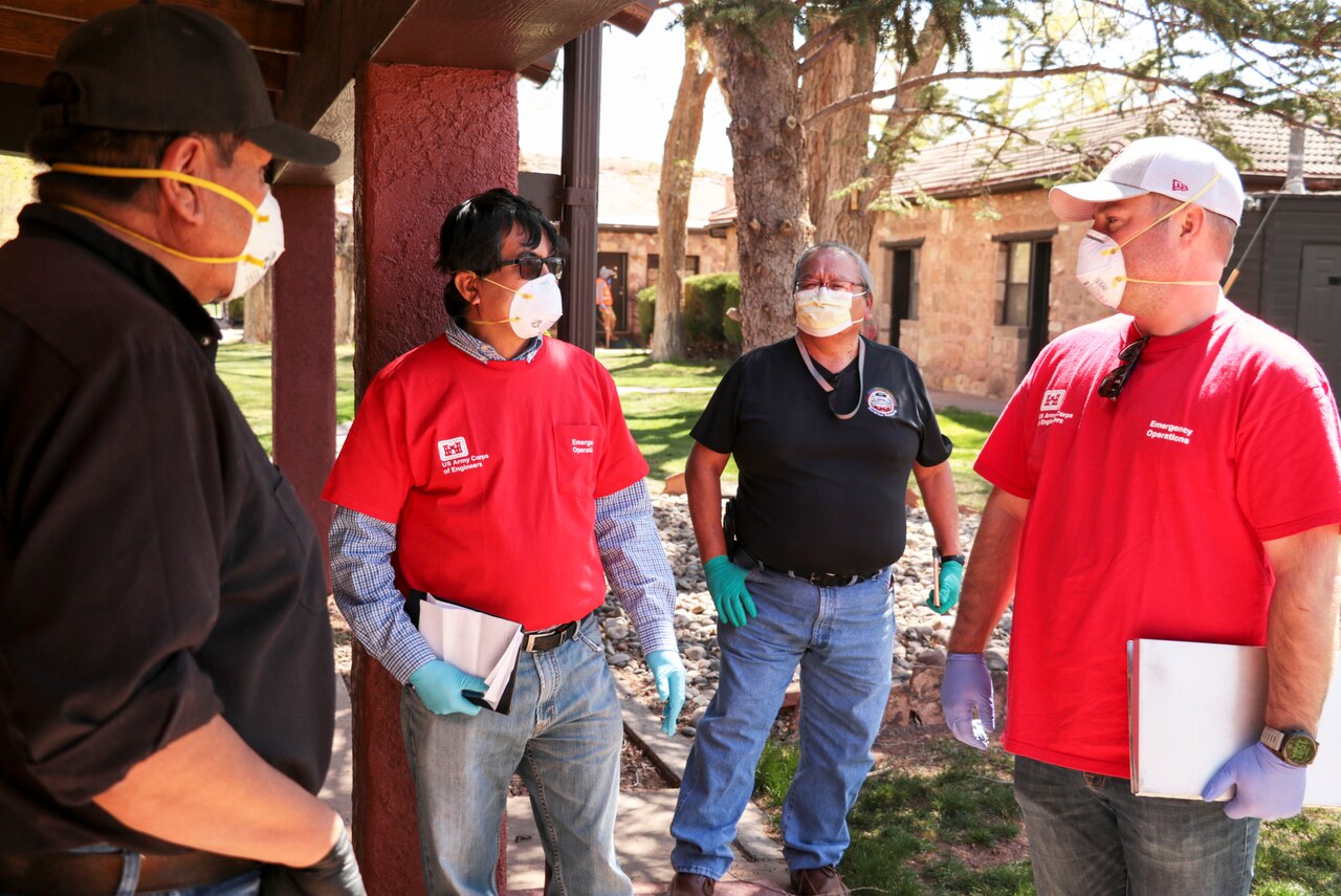 Four men in civilian clothing, wearing face masks and gloves, talk outdoors. There is a tree, a mound of dirt and buildings in the background.