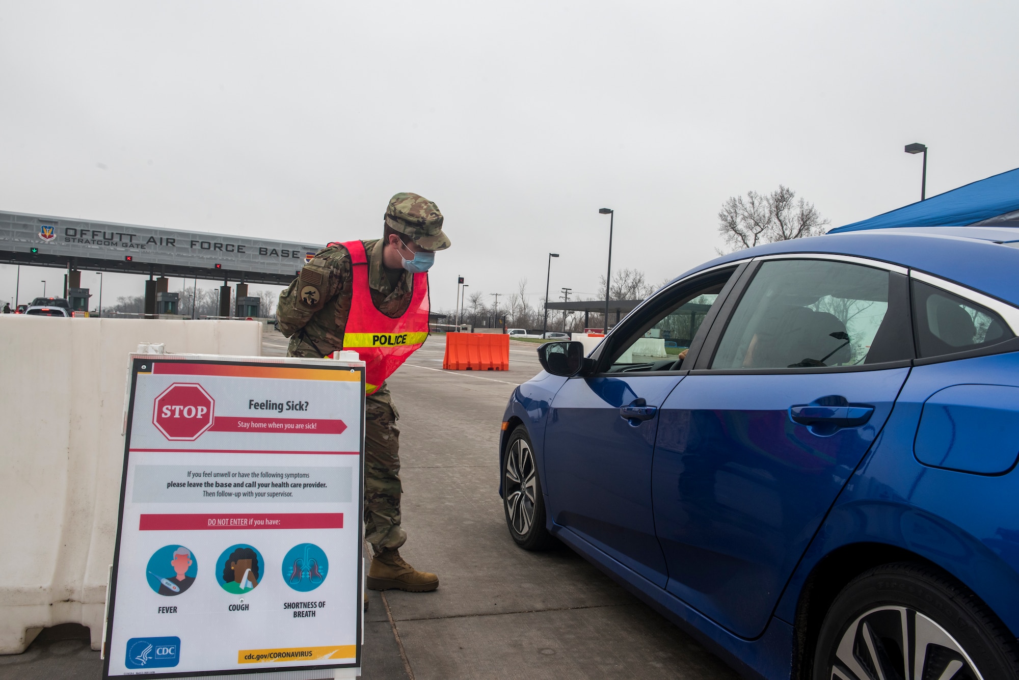A service member standing behind a COVID-19 symptoms sign bends down to look inside a car and ask the driver if they are experiencing any symptoms.