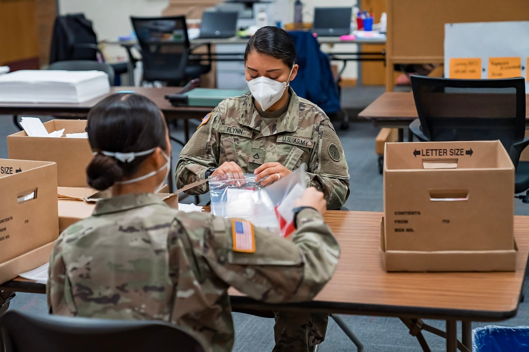 Two soldiers wearing facemasks sit across from each other and build test kits.