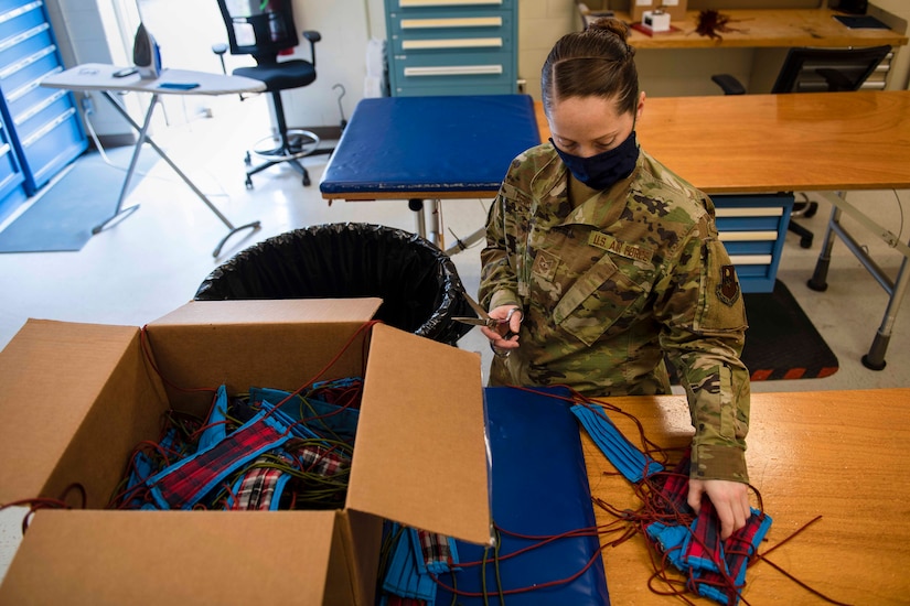 An airman wearing a facemask trims threads off facemasks on a table next to a large box.