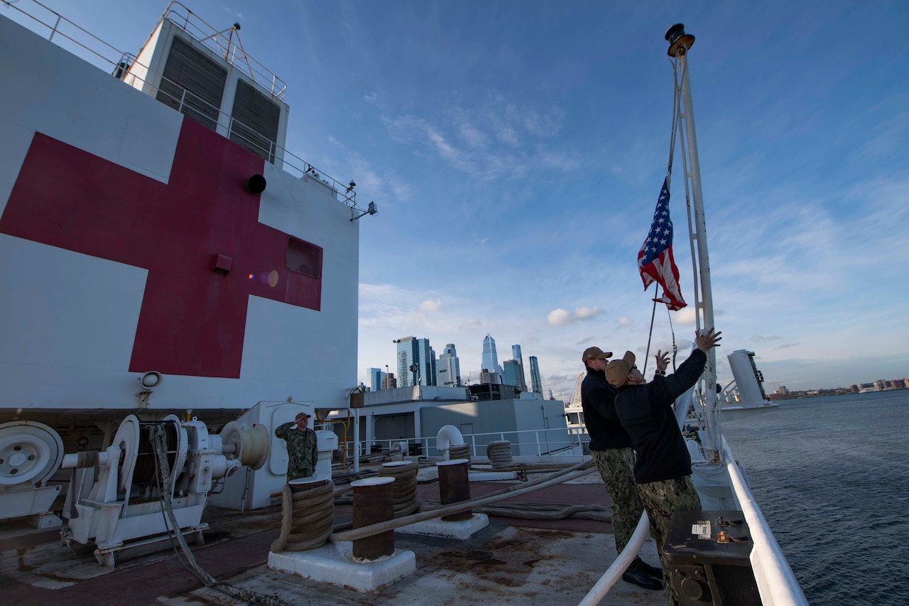Service members raise a flag aboard a military ship. The ship has a large red cross on a wall.