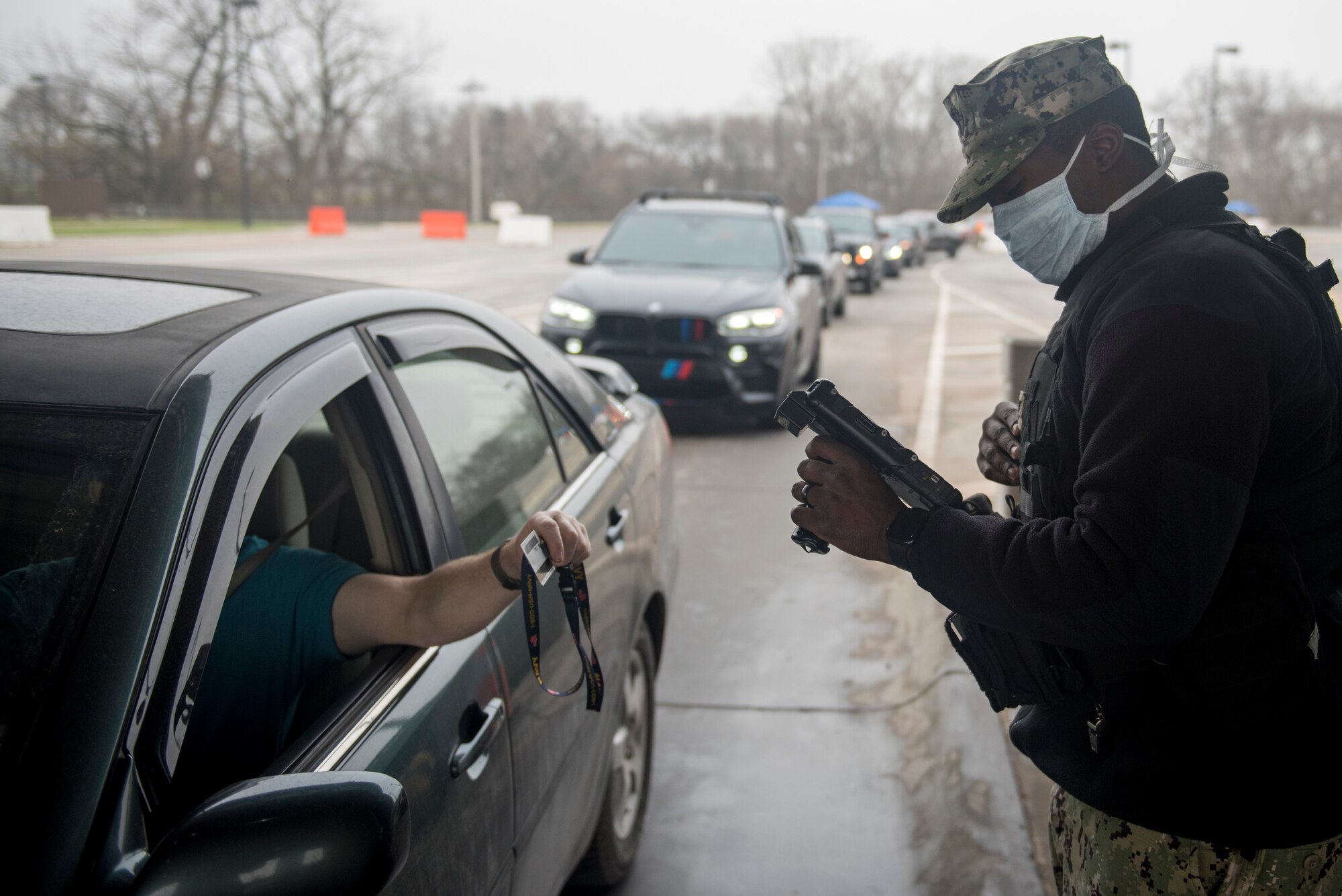 A service member wearing a cloth mask scans an ID card at an entry control point while a line of traffic waits to enter the installation.