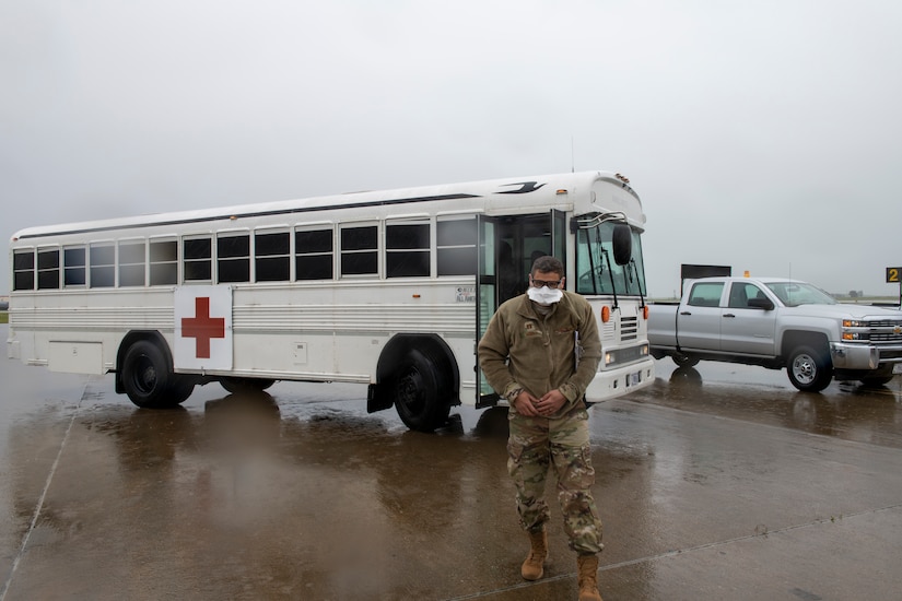 An airman wearing a white face mask walks away from a white bus marked with a red cross.