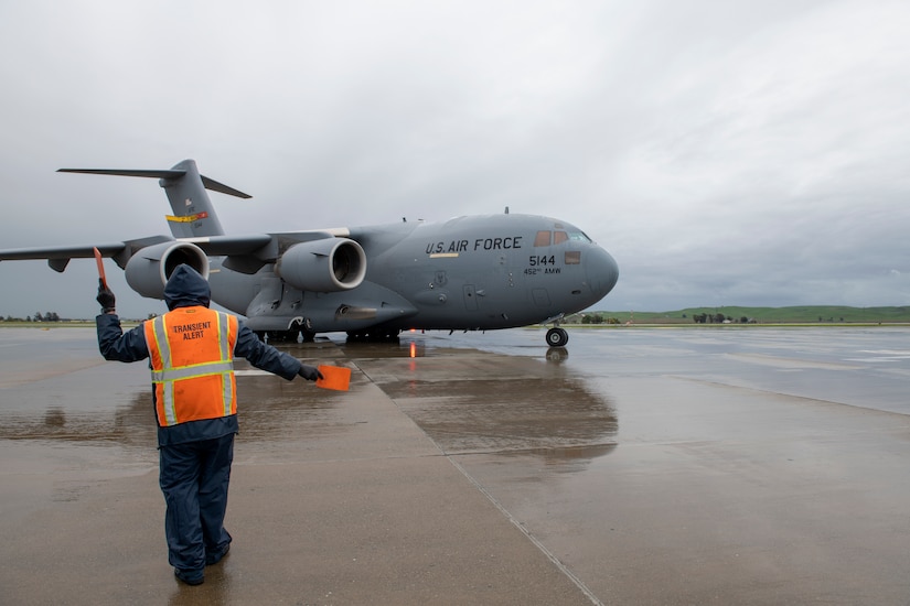 A man wearing blue coveralls and an orange vest marshals an aircraft into position.