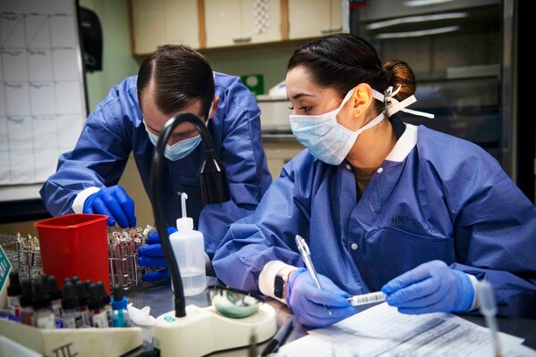 Two Navy medical technicians in personal protective equipment handle vials in a laboratory.