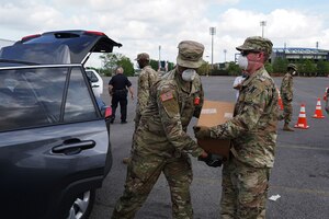 Two Guardsmen dressed in fatigues, and wearing protective face masks, unload boxes from a vehicle in a large parking lot.