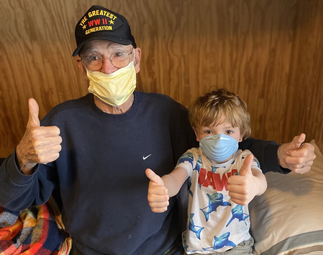 An elderly man and a small boy wearing medical masks give four thumbs up while sitting on a bed.