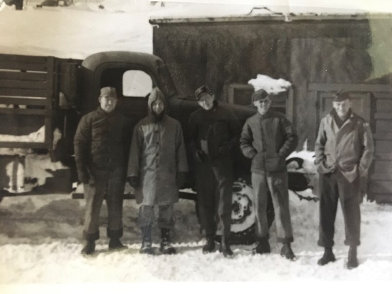 Five World War II Army soldiers stand in the snow in front of an old truck and a small building.