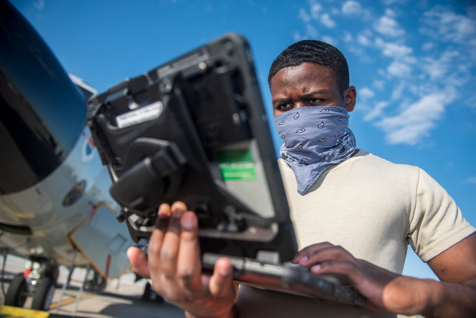 A servicemember reviews a technical order on a laptop as he prepares an aircraft for takeoff while donning a protective cloth mask.