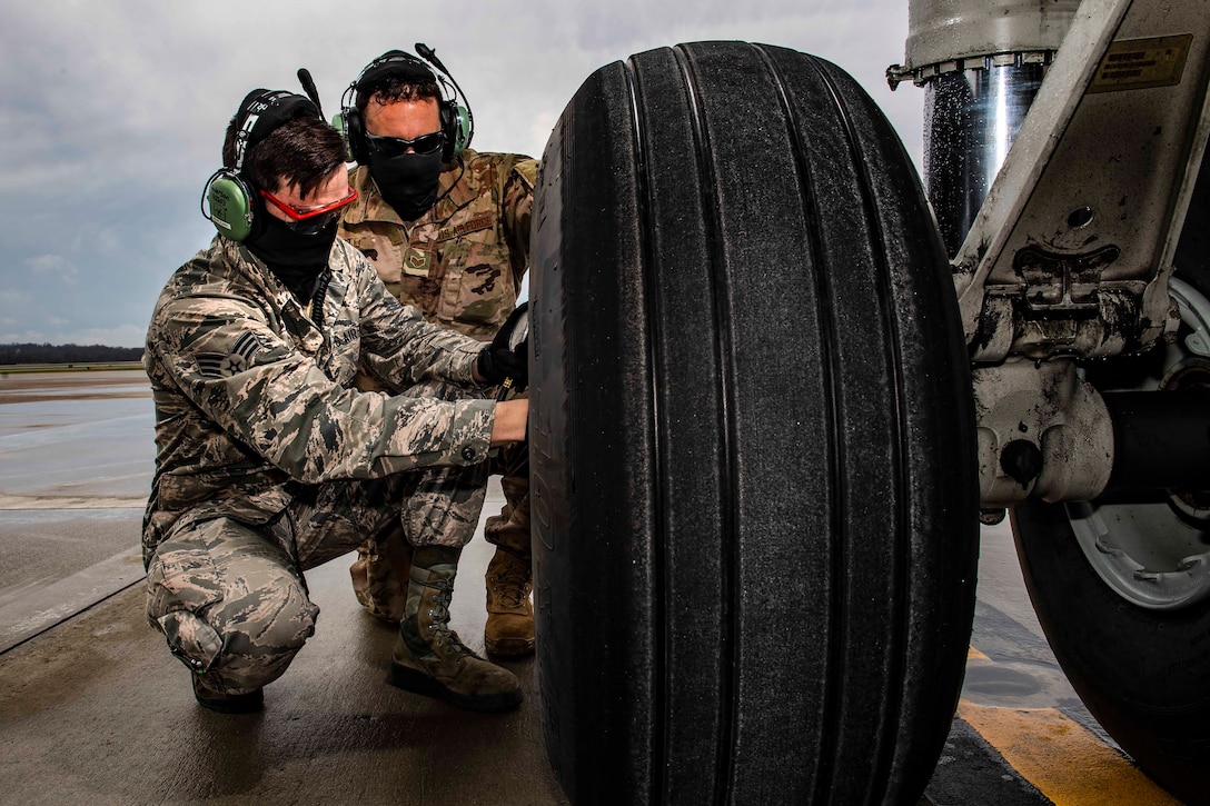 Two airmen wearing face masks kneel next to a large tire.