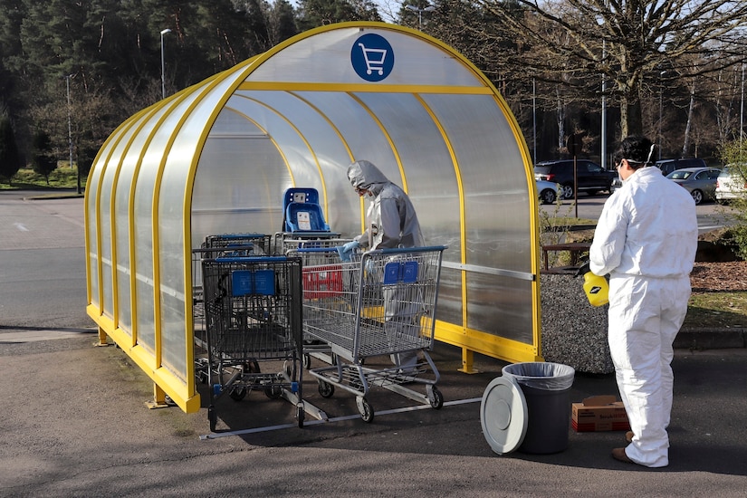 Two soldiers in white protective outfits work to clean grocery carts in a parking lot.