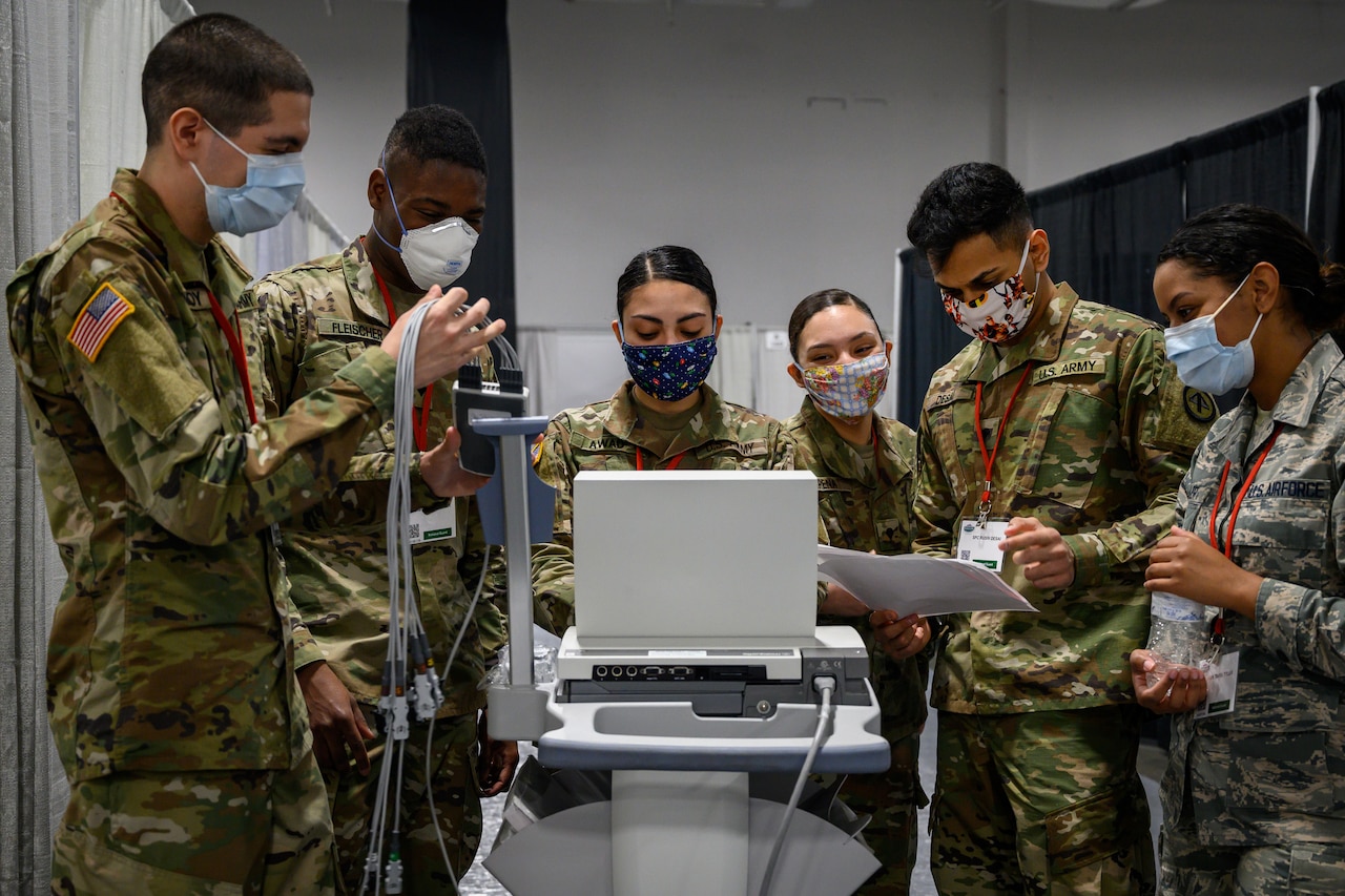 Soldiers and airmen in uniform and wearing face masks stand around an electrocardiogram machine.