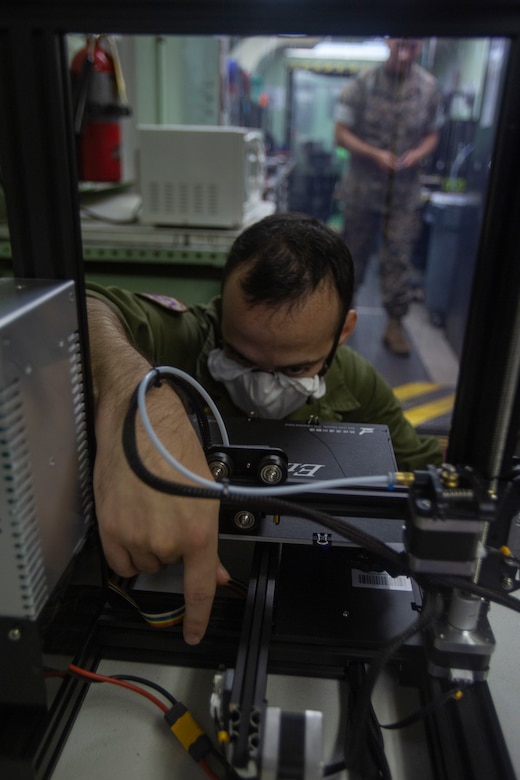 A Marine wearing a facemask performs maintenance on a 3D printer.
