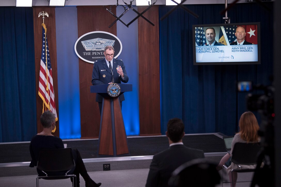 A service member speaks at a lectern to an audience of reporters sitting far apart from one another.
