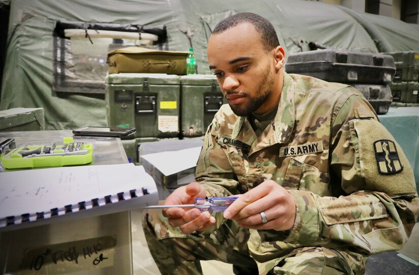 A service member crouches on the ground and uses a screwdriver on a piece of medical equipment.