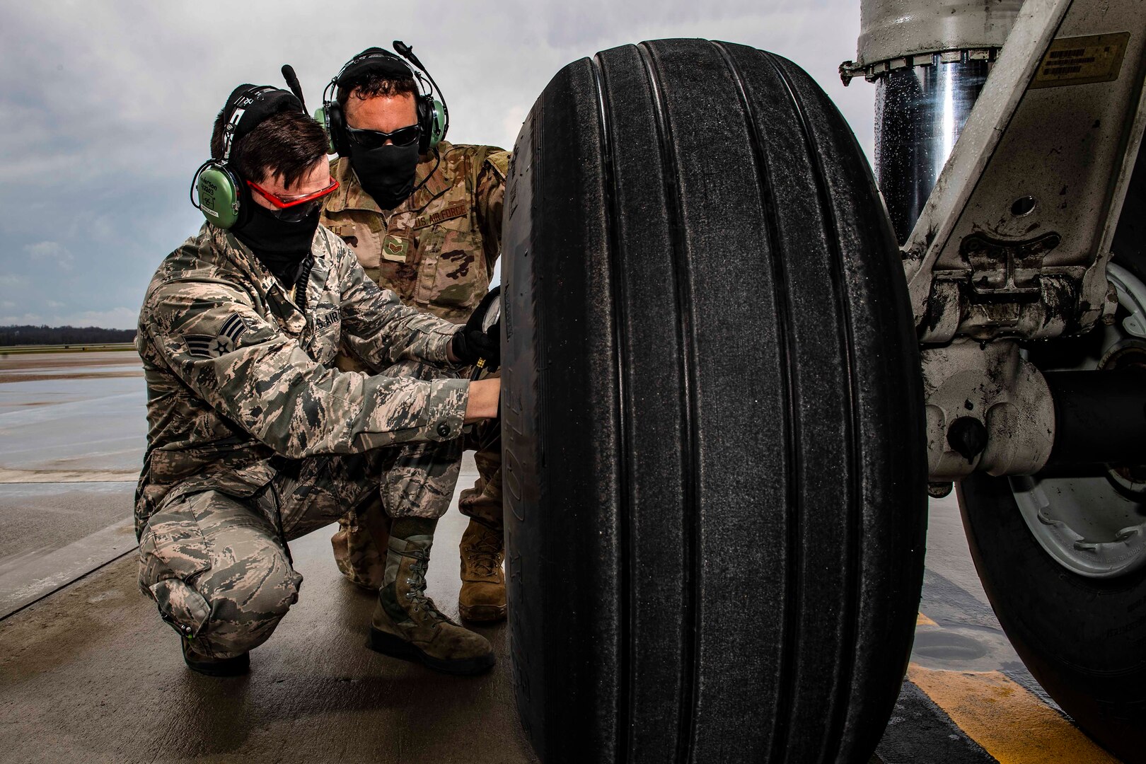 Two airmen wearing face masks kneel next to a large tire.