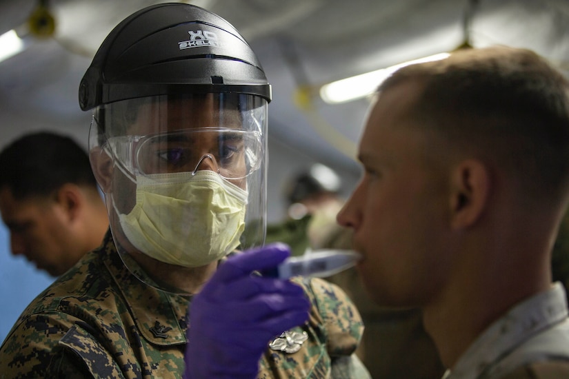 A sailor in protective gear takes a Marine’s temperature.