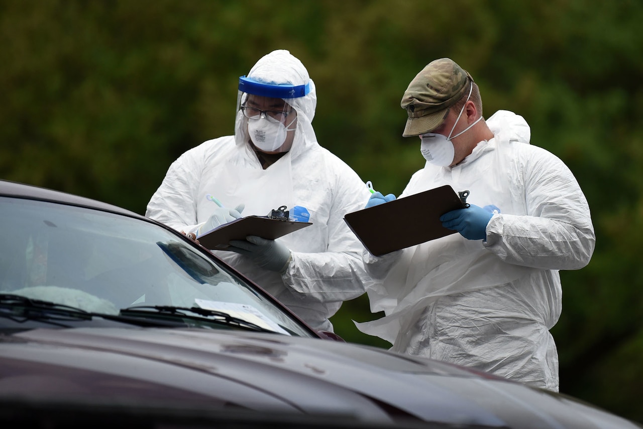 Two people in medical suits and masks hold clipboards.