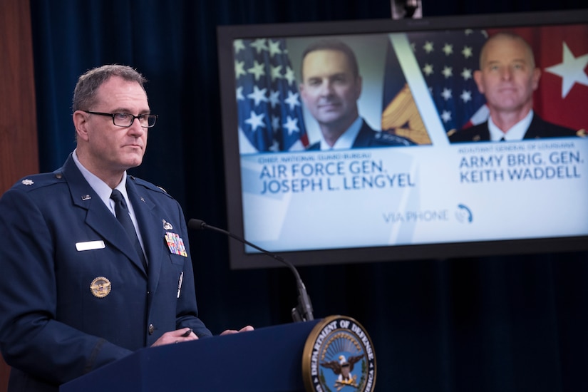 Man stands at lectern with large TV screen in background.