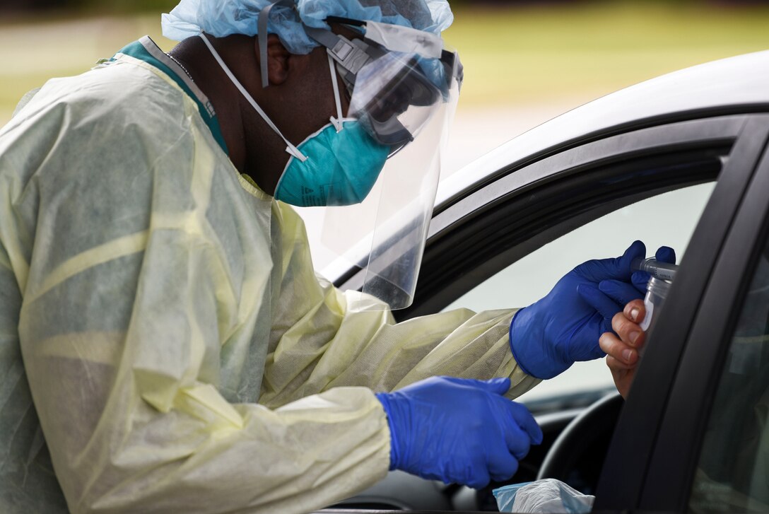 A person wearing personal protective equipment leans into a vehicle to administer a COVID-19 test.