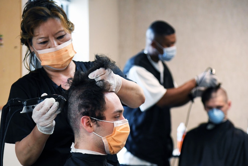 A female barber wearing personal protective equipment gives a new recruit the signature close-cropped haircut.