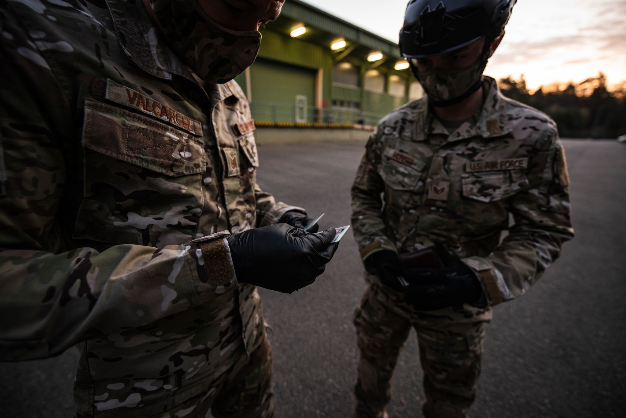 Two Airmen wearing gloves and cloth masks are verifying ID cards.