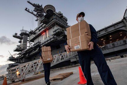 Two military personnel carry cardboard boxes. Both wear masks. In the background is an aircraft carrier.