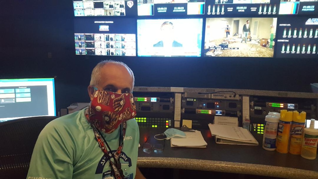 A man wearing a face mask sits at a desk with  technical equipment.