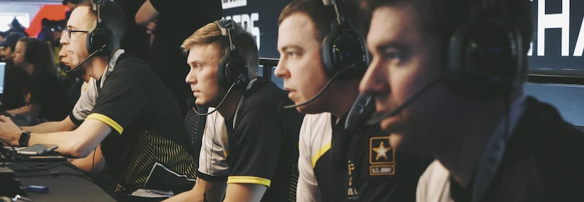 Four Soldiers in black and white jerseys with Army logos sit at computers with headsets on playing video games.