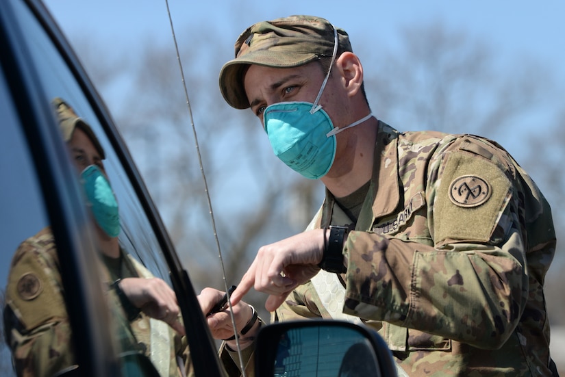 A soldier wearing a blue face mask checks a motorist’s identification.