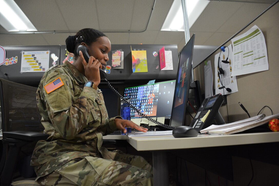 A soldier sitting at a desk speaks on a phone.
