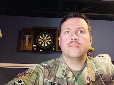 white male with mustache in green camouflage uniform with dark gray wall and dart board behind him.