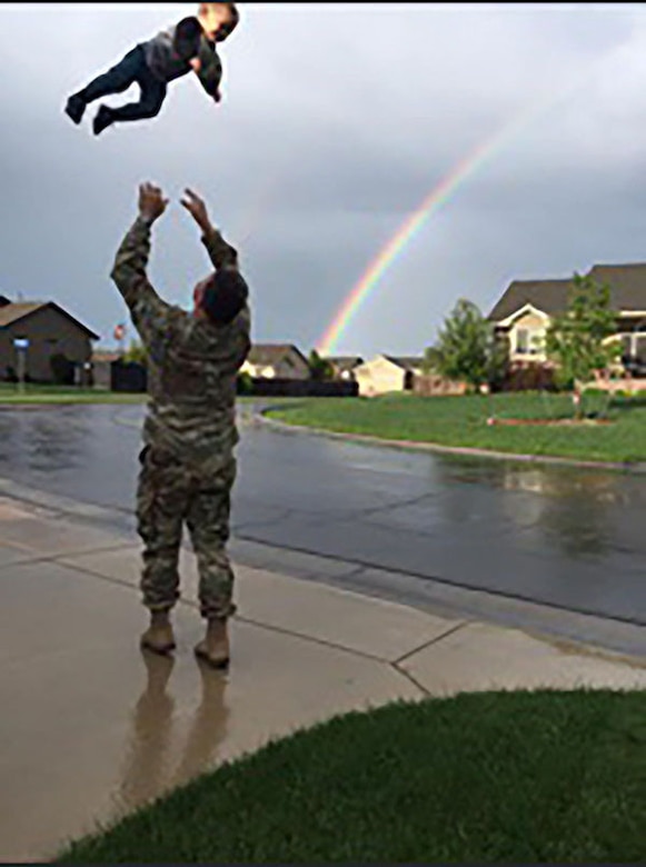 White man in green camouflage uniform and tan boots throw young boy in the air in a neighborhood. There is a rainbow in the sky in the background.