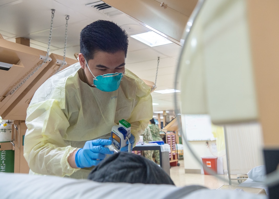 A sailor wearing a mask holds a thermometer near a patient's head.