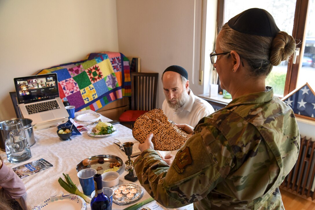 An Air Force chaplain breaks a piece of matzo at a table, as her family watches in person and others watch on a laptop screen.