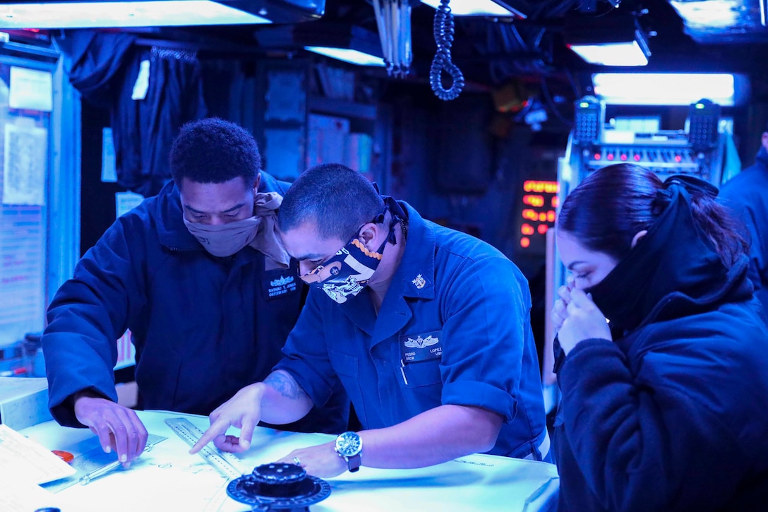 Three sailors wearing face masks look at papers on a table.