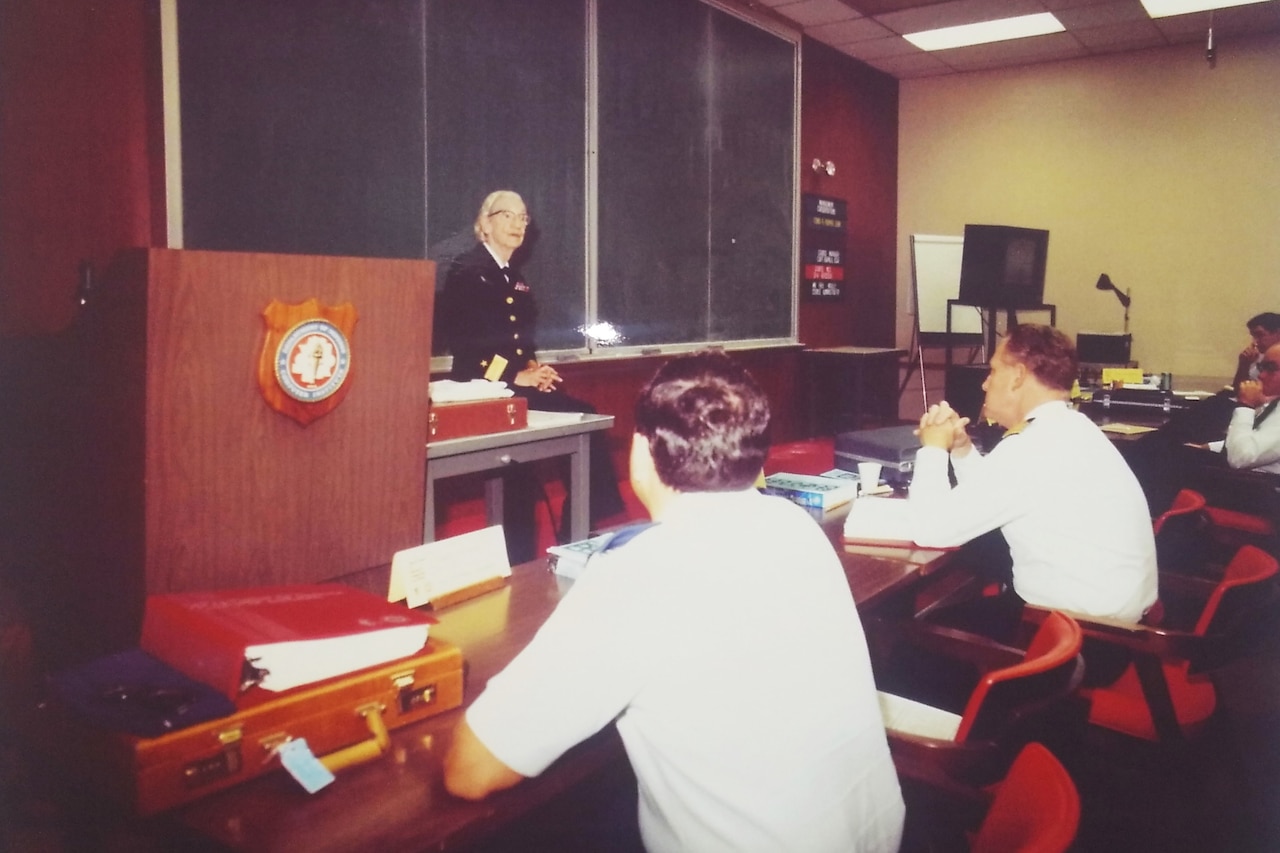 A woman in military uniform is seated at the front of a classroom.  Students seated at long tables listen.