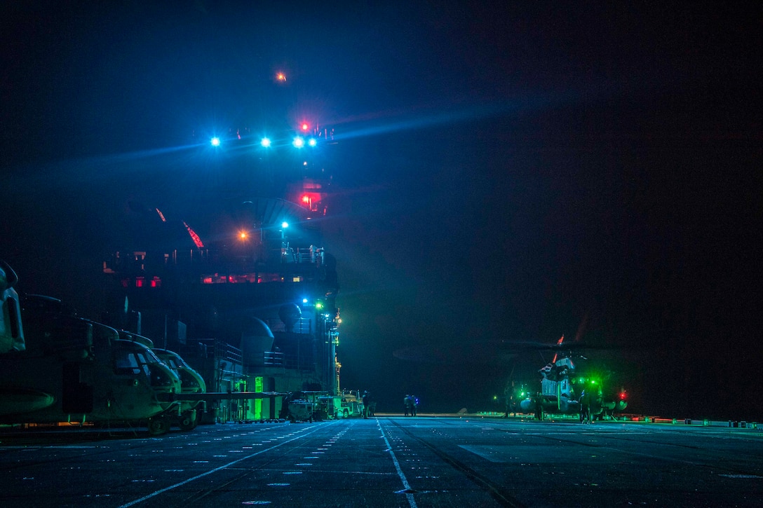 Service members stand next to aircraft on a ship’s deck at night.