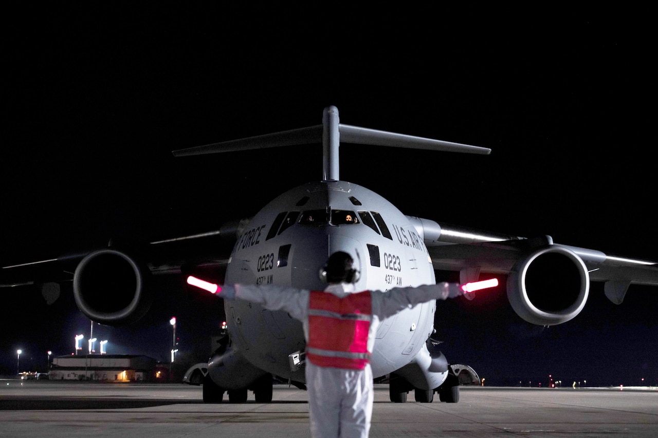 An airman holding out illuminated flashlights faces a large cargo jet as he guides it into position on a dark flight line.