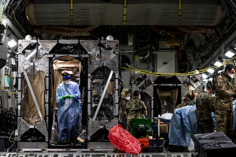 Airmen wearing personal protective gear work in and around an isolation chamber in the cargo bay of a large transport jet.